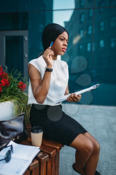 Business woman with notepad resting on the bench during the break in front of office building. Smiling black business woman in white blouse works outdoors