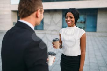 Business woman and businessman with coffee cups, outdoors meeting of partners, modern office building on background, partnership negotiations during the lunch break. Successful businesspeople