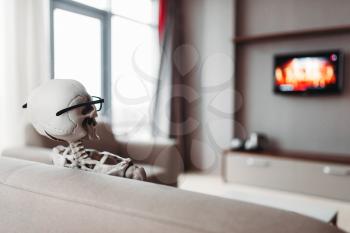 Skeleton in glasses is sitting on couch and watch tv, back view, window on background. Funny joke