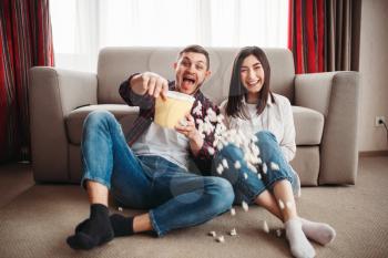 Laughing couple sitting on the floor against couch and watch comedy movie with popcorn at home, window on background