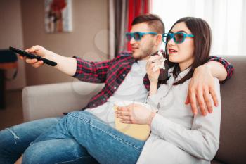 Smiling couple in 3D glasses sitting on couch and watch tv with popcorn at home, man with remote control in hand, window on background