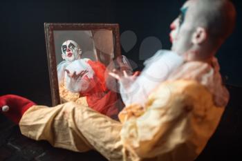 Portrait of scary bloody clown with crazy eyes sitting at the mirror, nightmare. Man with makeup in carnival costume, mad maniac
