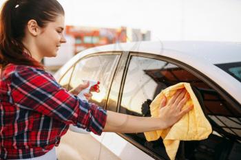 Cute woman cleans front glass of the car with sponge and spray, carwash. Lady on self-service automobile washing. Outdoor vehicle cleaning at summer day