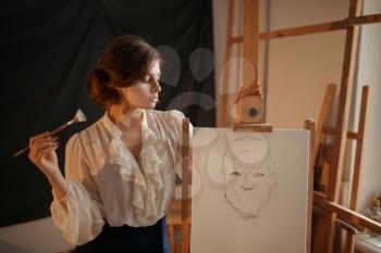 Cute female painter with brush standing against easel in studio. Creative paint, woman drawing pencil sketch, workshop interior on background