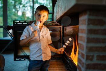 Little boy frying sausages on stick at fireplace in the house. Child cooking grilled meat on the fire right at home, happy childhood