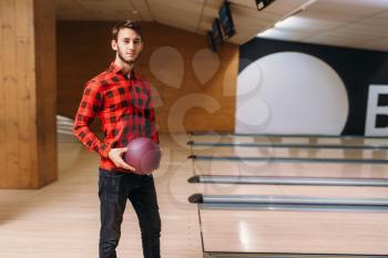 Male bowler standing on lane and holds ball in hands, back view. Bowling alley player poses in club, active leisure