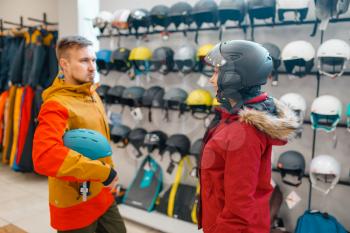 Young couple at the showcase trying on helmets for ski or snowboarding, side view, sports shop. Winter season extreme lifestyle, active leisure store, buyers looking on protect equipment