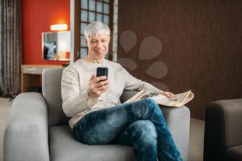 Adult man with phone and newspaper sitting on couch at home. Mature male person in jeans relax in armchair