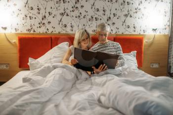 Mature couple looking at photo album in bedroom. Adult man and woman lying in bed before sleeping