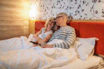 Mature love couple watching TV in bedroom before sleeping. Adult man in glasses and woman lying in bed
