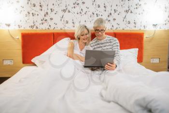 Mature couple looking on laptop in bedroom before sleeping. Adult man in glasses and woman lying in bed 