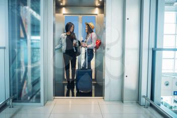 Two female travelers with luggage in airport elevator. Passengers with baggage in air terminal, back view, happy journey of white and black ladies, summer travel