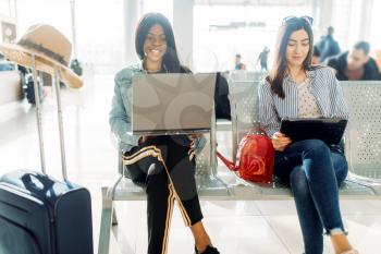 Two female travelers with laptops waiting for departure in airport. Passengers with baggage in air terminal, back view, happy journey of white and black ladies, summer travel