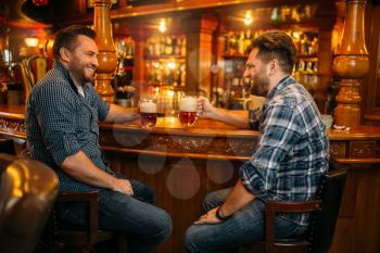 Two smiling male friends drinks beer at the counter in pub. Bearded men with mugs having fun in bar, good friendship