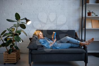 Young woman lying on cozy black couch and reading a book, living room in white tones on background. Attractive female person with magazine rest on sofa at home