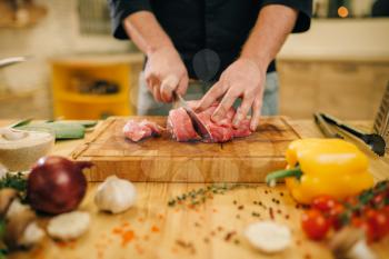 Male person with knife cuts raw meat into slices, closeup, kitchen interior on background. Chef cooking tenderloin with vegetables, spices and herbs