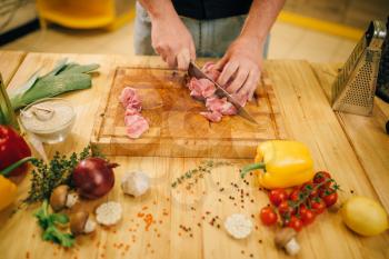 Male person hands with knife cuts raw meat into slices, top view, kitchen interior on background. Chef cooking tenderloin with vegetables, spices and herbs