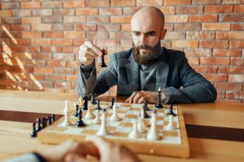 Male chess player playing black figures, queen move. Chessplayer at board, front view, intellectual tournament indoors. Chessboard on wooden table, strategy game