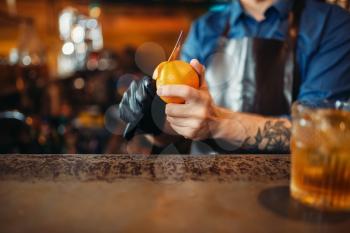 Male bartender in apron cleans orange at the bar counter. Alcohol beverage preparation. Barman occupation
