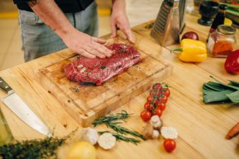 Male person hands seasoning piece of raw meat on wooden board, kitchen interior on background. Chef cooking tenderloin with vegetables, spices and herbs