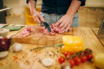 Male person hands with knife cuts raw meat into slices, top view, kitchen interior on background. Chef cooking tenderloin with vegetables