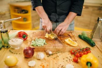 Chef hands with knife cuts yellow pepper on wooden board closeup. Man cutting vegetables, fresh salad cooking, kitchen interior on background. Male person chopping ingredients for lettuce
