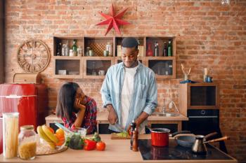 Black man cooking breakfast on the kitchen, wife drinks coffee. African couple preparing vegetable salad at home