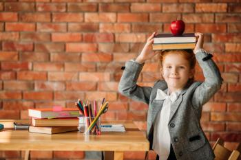 Cute schoolgirl sitting at the table, side view. Female pupil at the desk with textbooks, apples and globe, young girl doing homework