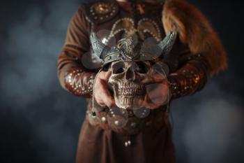 Viking dressed in traditional nordic clothes holds human skull in helmet, barbarian image. Ancient warrior in smoke on dark background