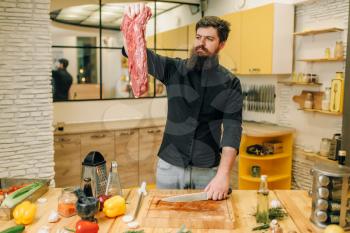 Man holds piece of fresh raw meat, kitchen interior on background. Chef cooking tenderloin with vegetables, spices and herbs