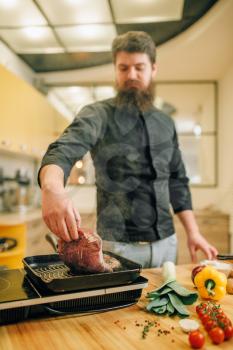 Bearded chef cooking meat in a pan on the kitchen. Man preparing boiled pork on table electric stove