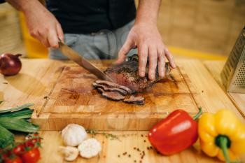 Male chef hands with knife cuts roasted meat on slices, top view. Man preparing beef with vegetables on countertop