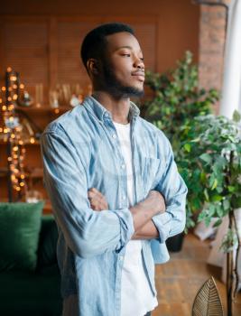 Black man with a cunning look in the living room, relaxation at home. Young african american male person standing in his house
