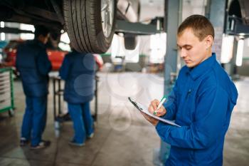 Repairman with notebook fills the check list, car on the lift, fixing the problems, suspension diagnostic. Automobile service, vehicle maintenance
