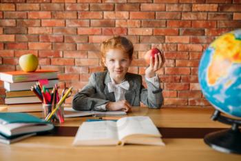 Funny schoolgirl with apple sitting at the table in classroom. Female pupil at the desk with textbooks and globe, school education
