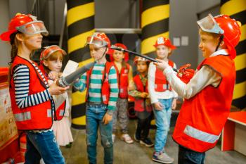 Children in helmet and uniform with hose and extinguisher in hands playing fireman, playroom indoor. Kids lerning firefighter profession. Child lifeguard, little heroes in equipment on playground