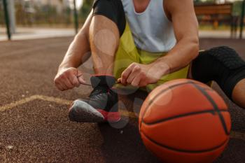 Basketball player sitting on the ground and tying laces on outdoor court. Male athlete in sportswear resting after streetball training