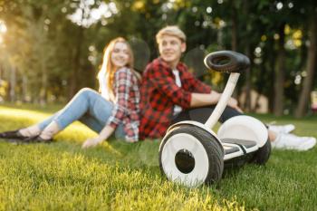 Young couple sitting on the grass near gyro board in summer park. Outdoor recreation with electric gyroboard. Eco transport with balance technology, electrical gyroscope vehicle