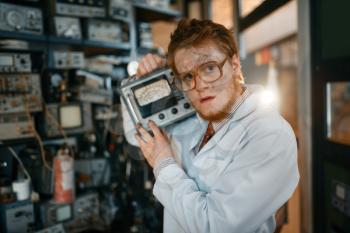 Crazy scientist in glasses holds electrical device in laboratory. Electrical testing tools on background. Lab equipment