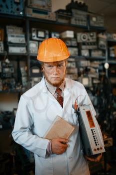 Strange male scientist in a helmet holding a book and an electronic device, engineer in laboratory. Electrical testing tools on background. Lab equipment, engineering workshop