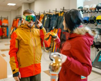 Couple at the showcase trying on mask for ski or snowboarding, shopping in sports shop. Winter season extreme lifestyle, active leisure store, customers buying glasses, protect equipment