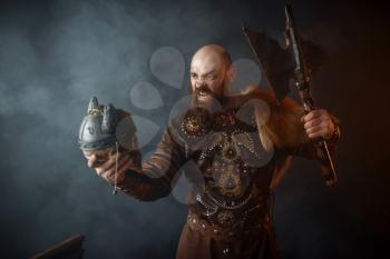Angry viking dressed in traditional nordic clothes holds enemy's skull in helmet and axe, barbarian image, murderer. Ancient warrior in smoke on dark background