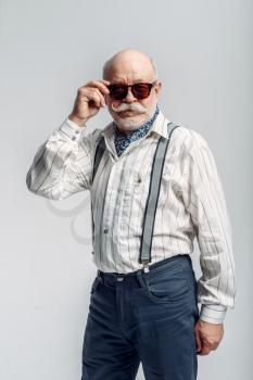 Bearded elderly man with mustache poses in sunglasses, grey background. Mature senior looking at camera in studio