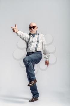 Fashionable elderly man makes fingers like a gun, grey background. Mature senior looking at camera in studio, dude