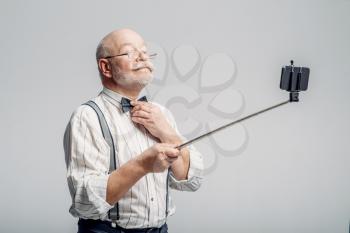 Happy elderly man makes selfie on phone, grey background. Mature senior showing his tongues at camera in studio