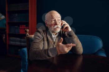 Elderly man talking by mobile phone in home office. Bearded mature senior in living room, old age businessman