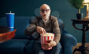 Elderly man with popcorn watching TV on couch in home office. Bearded mature senior poses in living room, old age businessman