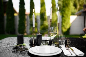 Table setting, glasses, candles and plate on black top, nobody. Luxury silverware, tableware outdoors, elegant decoration. Romantic celebration on summer meadow