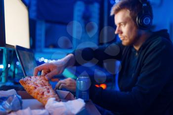 Male gamer eating pizza at his workplace with laptop and desktop PC, gaming lifestyle, night tournament. Computer games player in his room with neon light, streamer