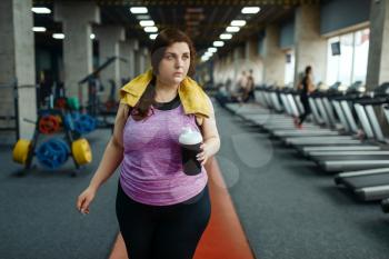 Overweight woman drinks diet cocktail in gym, active training. Obese female person struggles with excess weight, aerobic workout against obesity, sport club